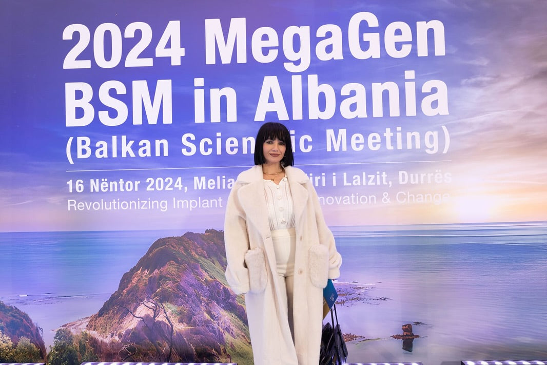 Woman standing in front of a promotional banner for 2024 MegaGen BSM in Albania with scenic background.