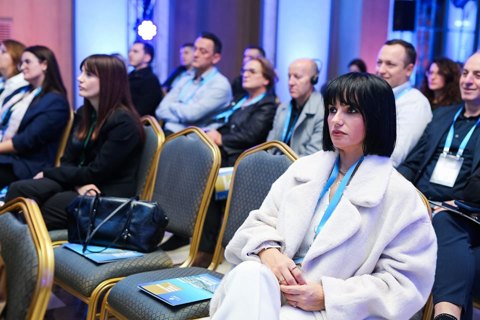 Woman in a white coat sitting in a conference room with other attendees.