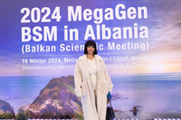 Woman standing in front of a promotional banner for 2024 MegaGen BSM in Albania with scenic background.